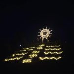 Kalno šlaitas su šviesų instaliacija, vaizduojančia saulę ir ornamentus tamsiame fone.